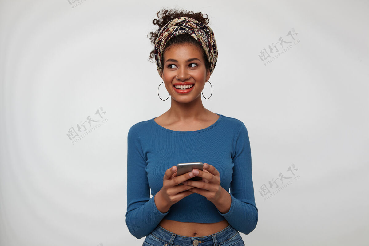 姿势摄影棚拍摄的年轻快乐美丽卷曲的深褐色女性微笑着广泛的一边看 保持手机在举起的手 而在白色的背景上摆姿势微笑年轻鼻子