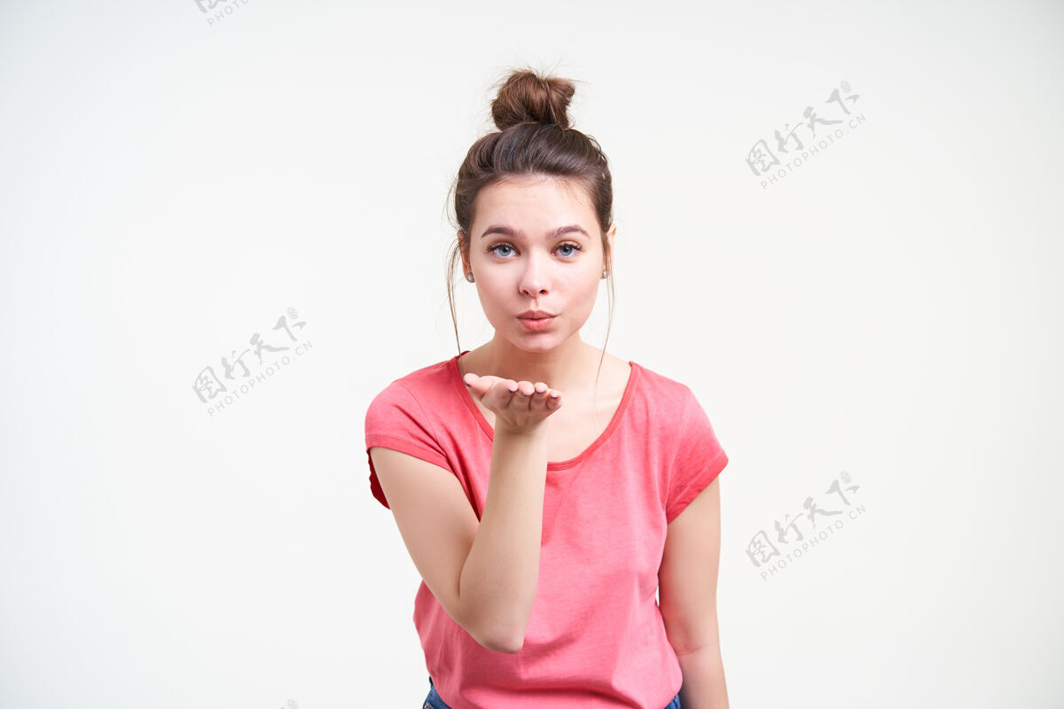 皮肤白皙摄影棚拍摄的一张年轻迷人的棕色头发女士举着手掌对着镜头吹气亲吻 穿着粉色t恤站在白色背景上表情空气休闲