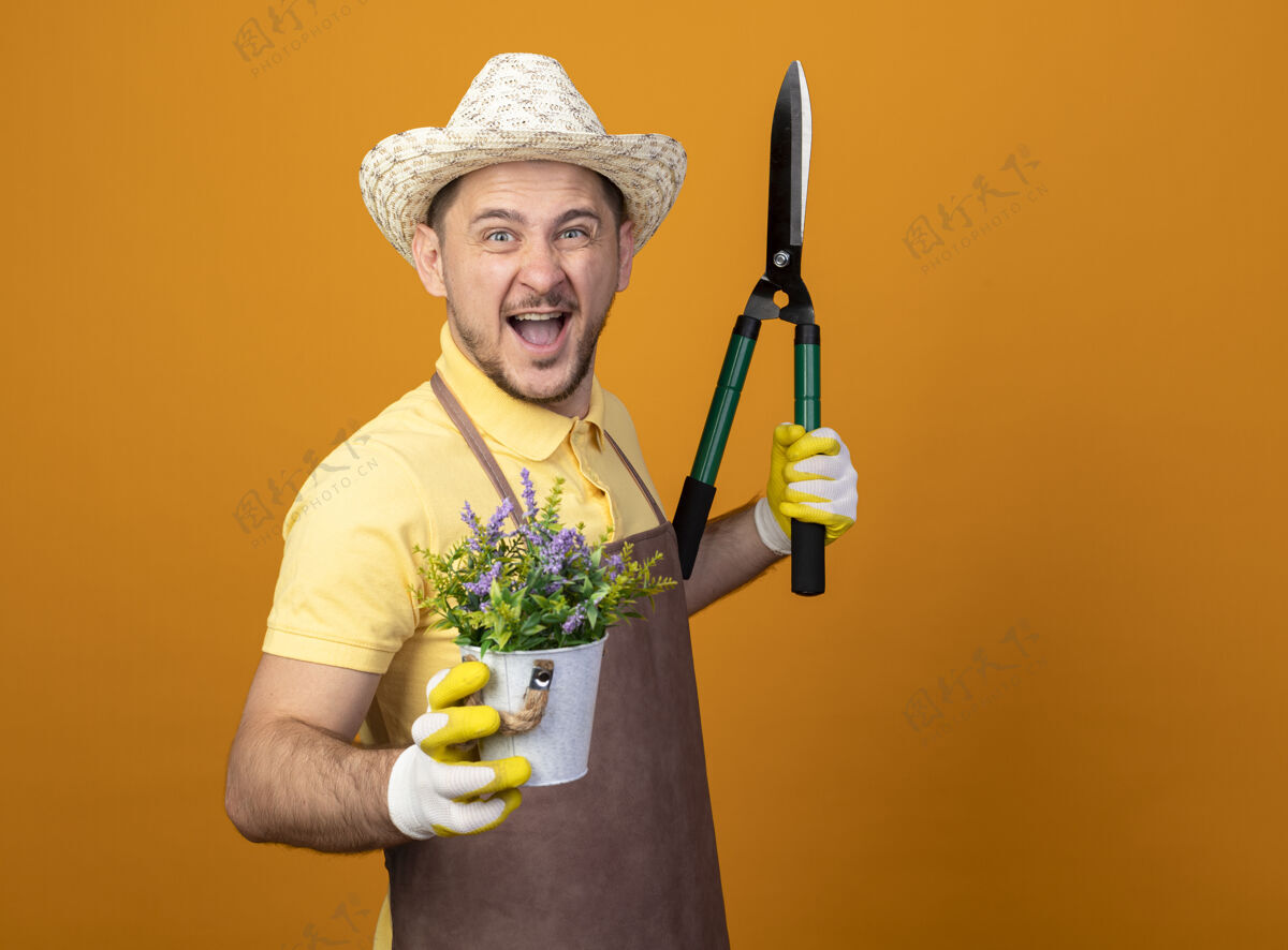 兴奋快乐的年轻园丁 穿着连体衣 戴着帽子 拿着树篱剪和盆栽植物 快乐而兴奋地笑着剪子穿植物