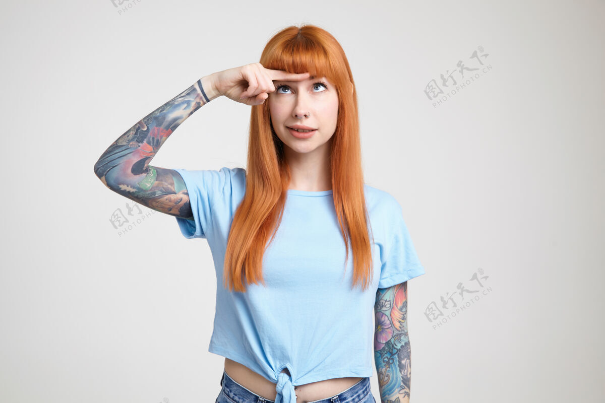 长迷惑不解的年轻漂亮的纹身女子 留着一头蓬松的长发 把食指放在额头上 一边看着它 一边穿着蓝色t恤站在白色背景上表情白皙食指