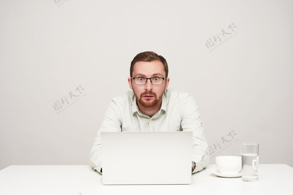 休闲戴眼镜的年轻短发胡须男子激动不已 双手放在笔记本电脑的键盘上 惊讶地看着摄像机 被隔离在白色背景下手表男人笔记本电脑