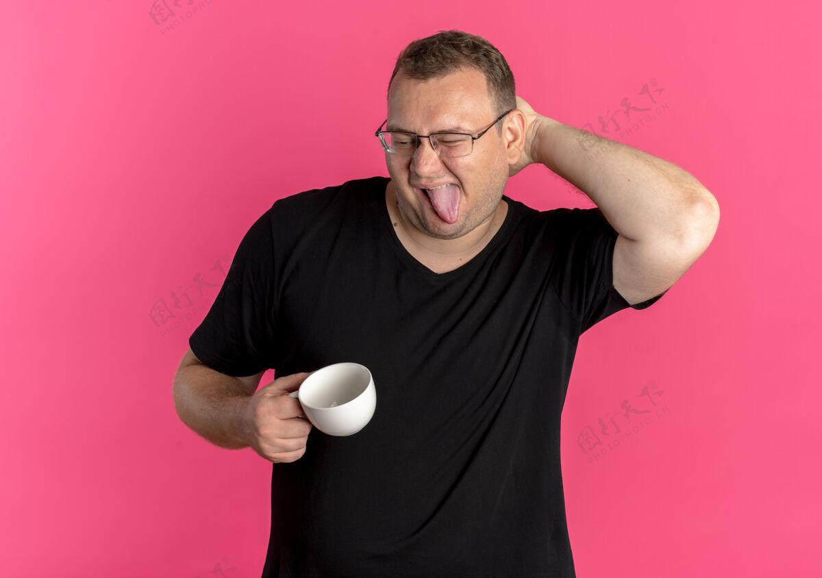粘一个戴眼镜的胖男人穿着黑色t恤 手里拿着咖啡杯 正开心地伸出舌头 站在粉红色的墙上挠头超重乐趣杯子
