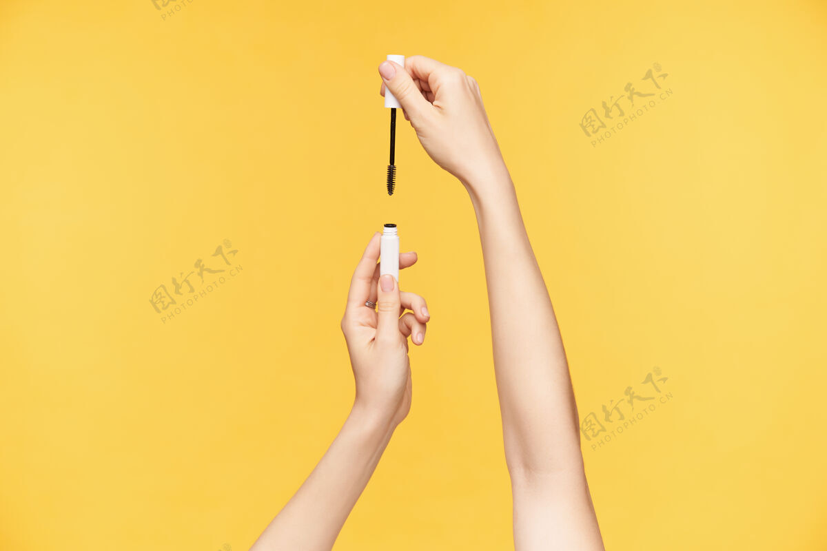白皙室内拍摄的年轻漂亮女人的手与裸体美甲被提出 而持有睫毛膏瓶 要染色睫毛 而被隔离在橙色背景墨水刷子手指