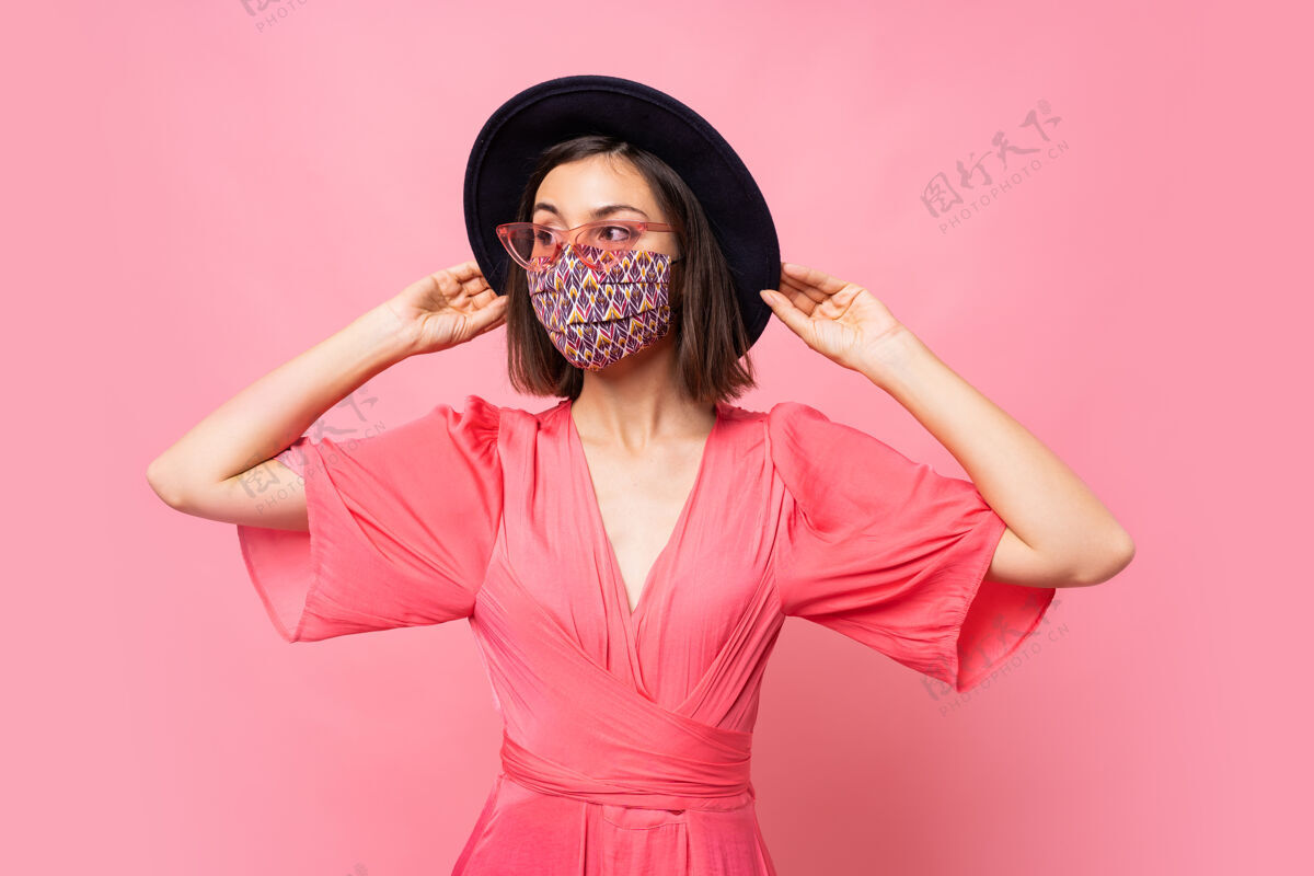距离时髦的女人戴着保护性的时髦的面具戴着黑色的帽子和太阳镜在粉红色的墙上摆姿势黑发社交休闲