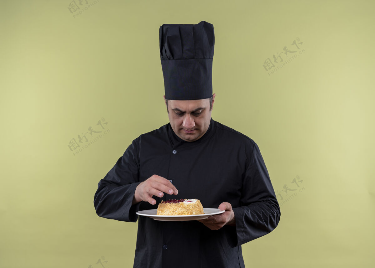 看男厨师身穿黑色制服 头戴厨师帽 手拿蛋糕盘 严肃的脸站在绿色背景上看着它厨师盘子黑色
