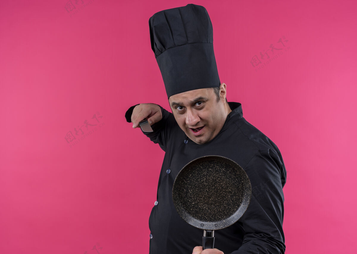 平底锅男厨师身穿黑色制服 头戴厨师帽 拿着平底锅和刀 严肃的脸站在粉色背景下看着镜头拿着制服穿