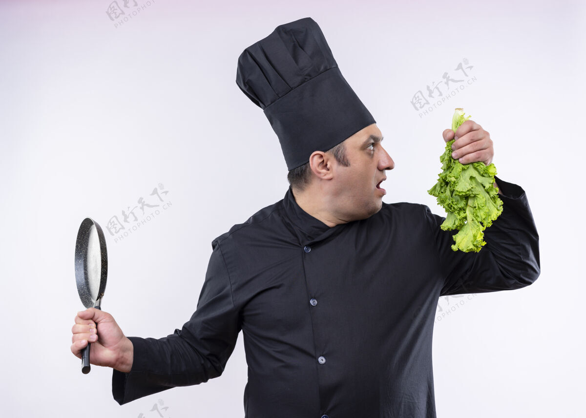 站着男厨师身穿黑色制服 头戴厨师帽 手里拿着新鲜的莴苣和平底锅 站在白色背景上惊讶地看着一旁帽子平底锅黑