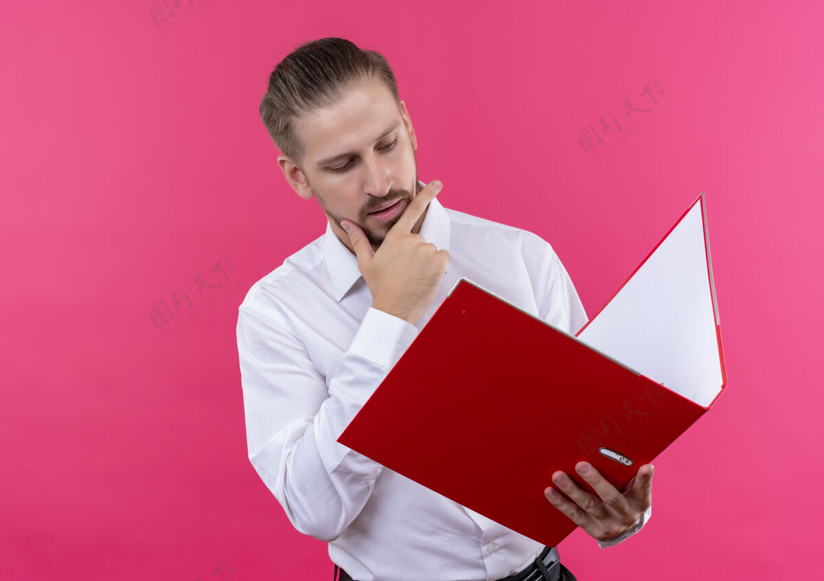 粉色穿着白衬衫的帅哥商人拿着文件夹看着它 严肃的脸站在粉色的背景上抱着帅气站着