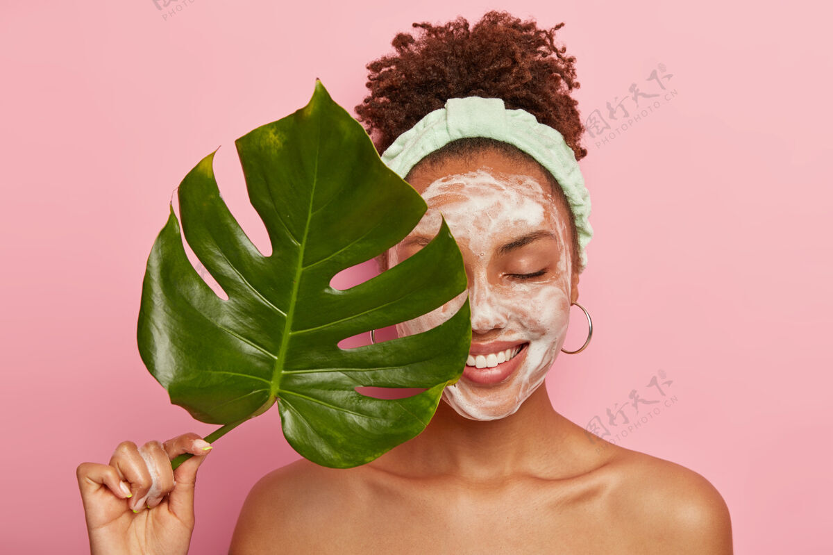 黑发快乐的非洲裔美国女人的画像用绿叶遮住半张脸 清洁脸部 用肥皂泡洗 赤裸上身站立 关心她的美丽和身体绿色牙齿泡沫