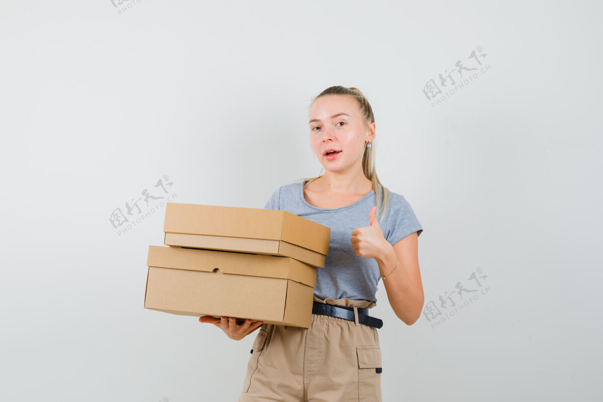 时尚年轻女性拿着纸板箱 穿着t恤 裤子 大拇指朝上 看起来很高兴正面图显示休闲交付