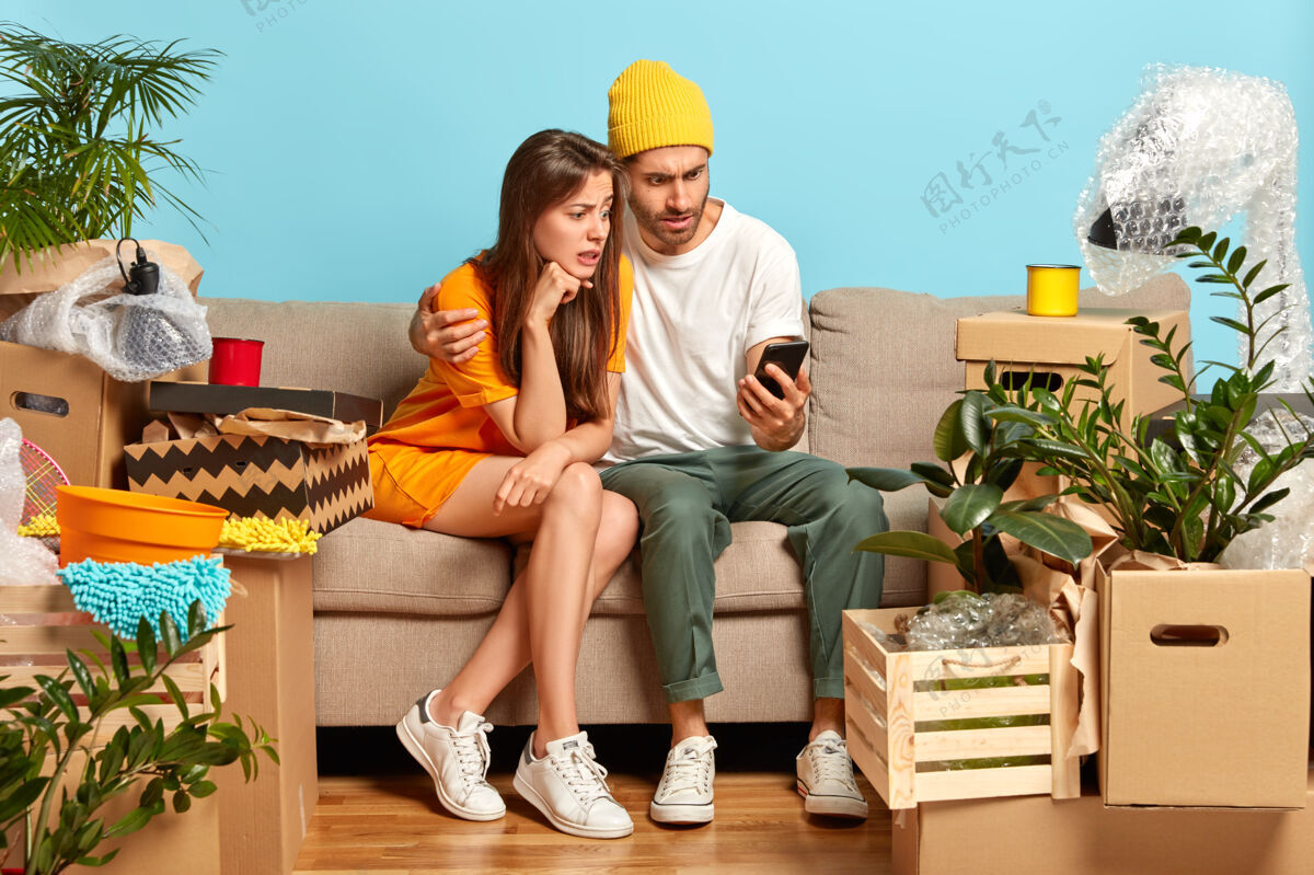 手机困惑的女人和男人看着智能手机设备 搬进新公寓 在网上商店寻找家具打开包装包装女性