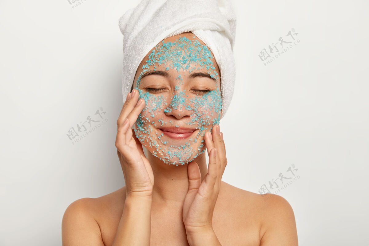 年轻化年轻开朗的女人在脸上涂上天然的磨砂膏 抚摸脸颊 闭上眼睛 戴上毛巾 淋浴后做美容 在室内做模特女模特皮肤上涂上蓝色海盐微笑卫生治疗