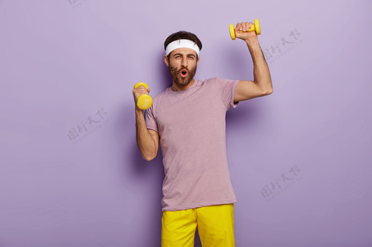 情绪有趣的男人有乐趣 练习哑铃 穿着运动服 为健康的生活方式而努力 早上有规律的训练紫色有趣头带