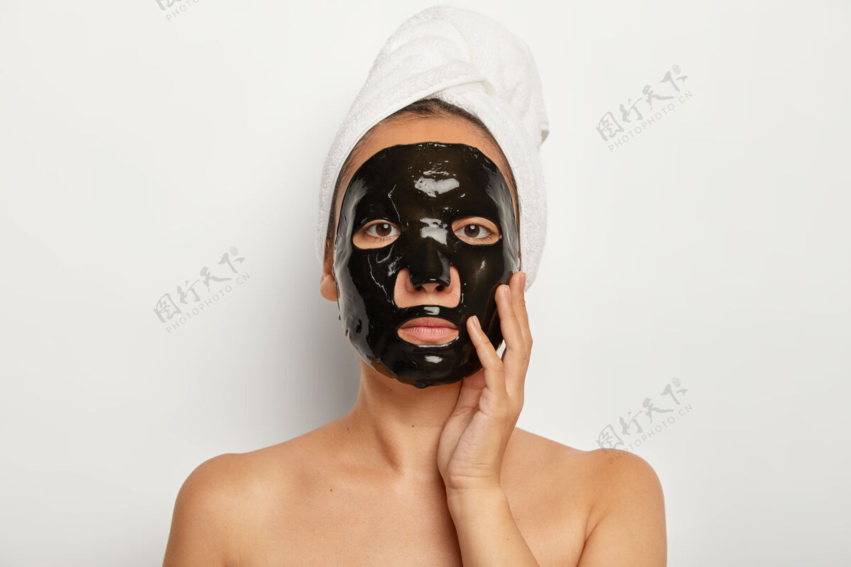 健康认真的亚洲女性在家里有美容程序 应用黑色净化面膜 直视 轻轻抚摸脸颊 头上戴着白色软毛巾年轻健康闭嘴
