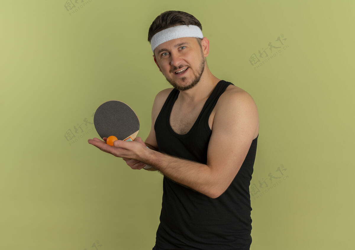 球拍戴着头巾拿着球拍和乒乓球的年轻健身男子站在橄榄色的背景下微笑着去打乒乓球乒乓球微笑橄榄