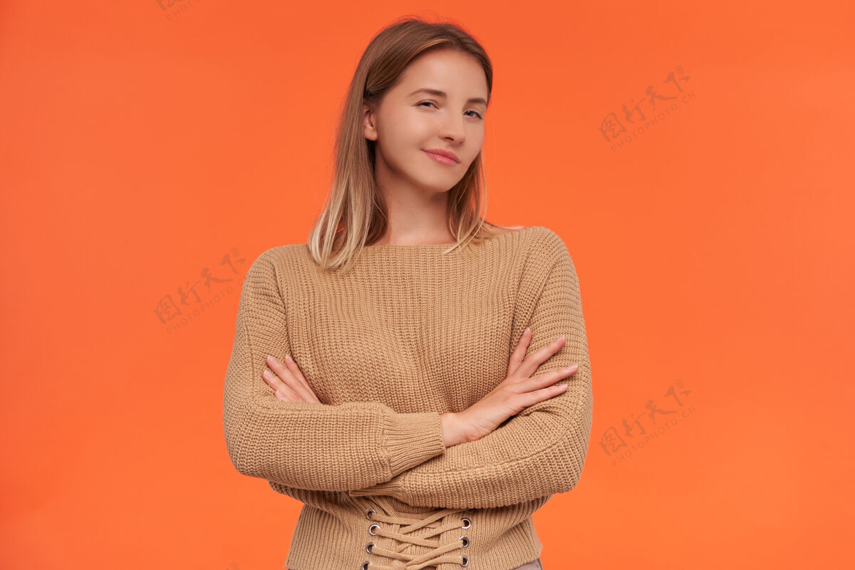 宽松室内拍摄的年轻迷人的女子穿着米色毛衣 双手叠在胸前 在橙色的墙上摆姿势 眯着眼睛 狡猾地看着前面爱情斜视表情