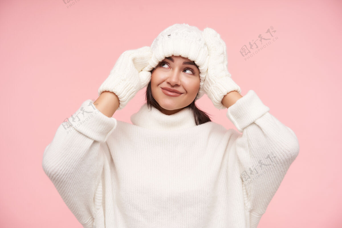 表情室内拍摄的年轻漂亮的棕色头发的女人触摸她的白色帽子 举起双手 微微微笑 穿着舒适的衣服站在粉红色的墙上女人提高自然