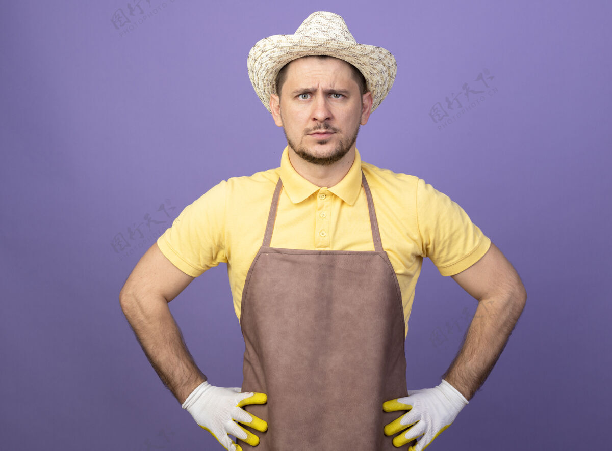 工人年轻的园丁穿着连体衣戴着帽子戴着工作手套站在紫色的墙上严肃地看着前面人工作手套