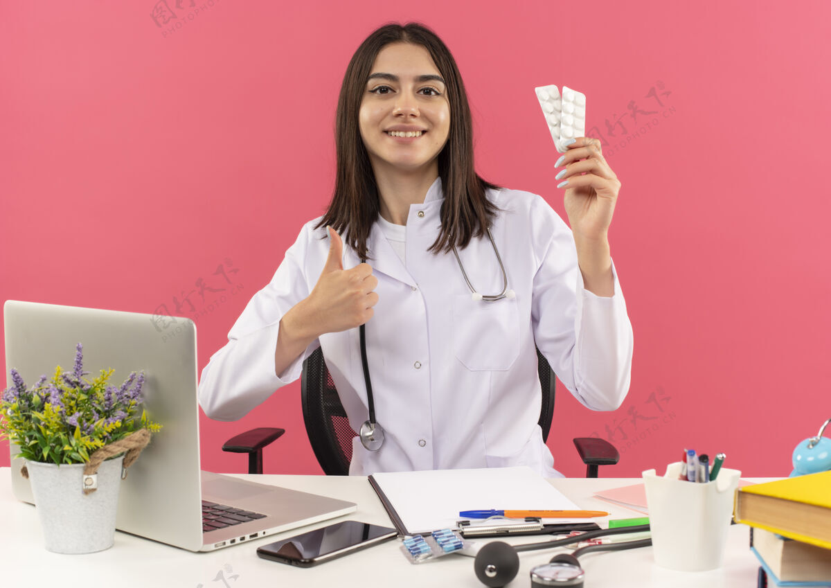 市民年轻的女医生穿着白大褂 脖子上戴着听诊器 手里拿着水泡 手里拿着药片 大拇指朝上 微笑着坐在桌旁 笔记本电脑放在粉色的墙上听诊器笔记本电脑桌子
