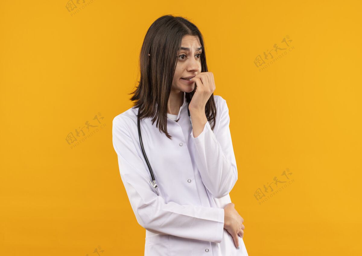 姿势年轻的女医生穿着白大褂 脖子上戴着听诊器 站在橙色的墙上 紧张地看着一边看外套手势