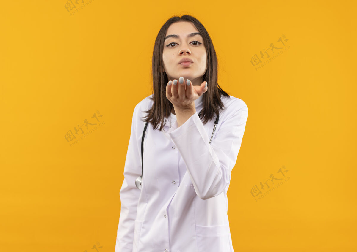 女性年轻的女医生穿着白大褂 脖子上戴着听诊器 站在橘色的墙上 用手在她面前吹吻吻医生吹