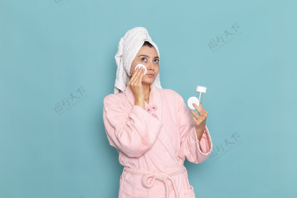 化妆品正面图身着粉色浴袍的年轻女性在蓝色墙壁上洗脸化妆清洁美丽净水自我护理霜淋浴前面年轻人奶油