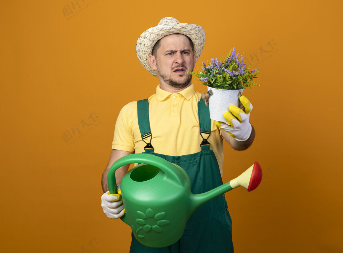 市民年轻的园丁穿着连体衣 戴着帽子 手里拿着浇水罐 站在橘红色的墙上 看着手中的盆栽植物 既不高兴又困惑站着制服植物