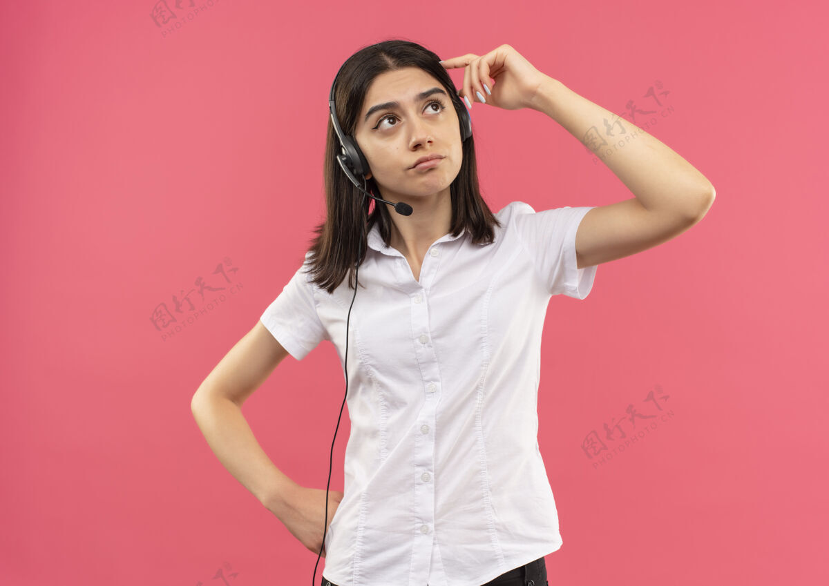 人穿着白衬衫戴着耳机的年轻女孩 站在粉红色的墙上困惑地抬头看拼图人手势