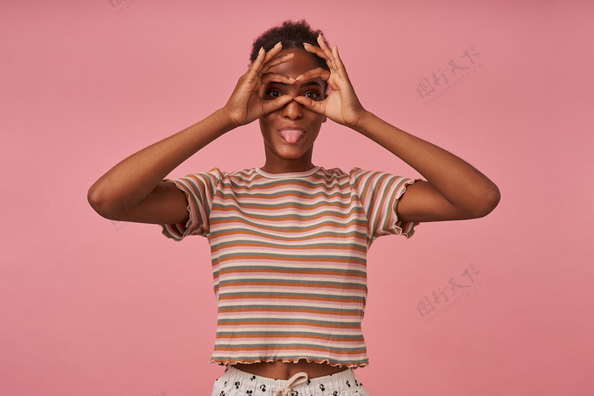 表情室内拍摄的年轻黑发女性戴着眼镜 穿着条纹t恤站在粉色墙壁上 双手合十 露出欢快的舌头姿势女士心情