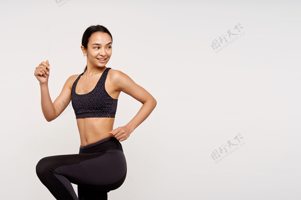 女人积极的年轻迷人的运动型黑发女子 没有化妆 举手踱步 愉快地看着身后 隔着白墙健身训练运动装