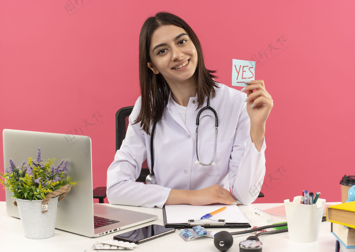 年轻人年轻的女医生穿着白大褂 脖子上戴着听诊器 手里拿着写着“是”的提示纸 面带微笑 坐在桌旁 笔记本电脑放在粉红色的墙上听诊器医院微笑