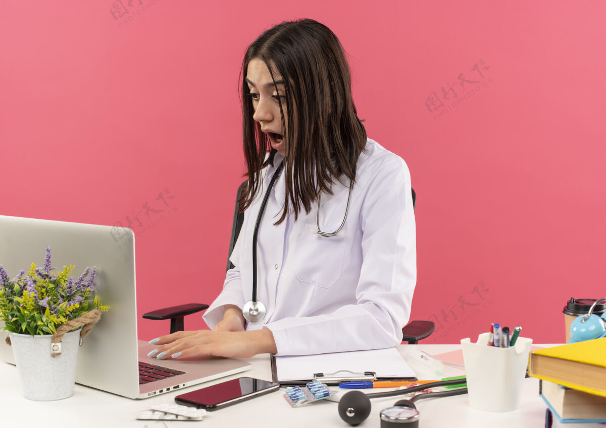 外套年轻的女医生穿着白大褂 脖子上戴着听诊器 坐在桌旁 手提电脑在粉红色的墙上显得困惑和惊讶坐着看医疗保健