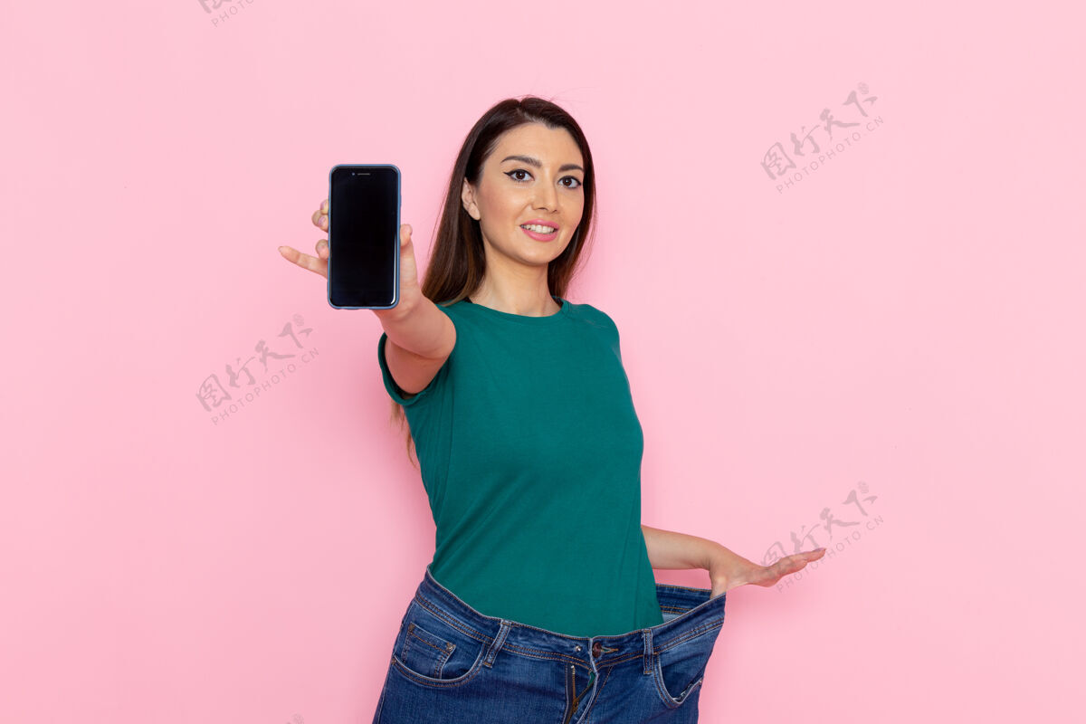 运动正面图身着绿色t恤的年轻女性手持智能手机在浅粉色墙壁上腰部运动锻炼美丽苗条女性运动减肥腰部运动