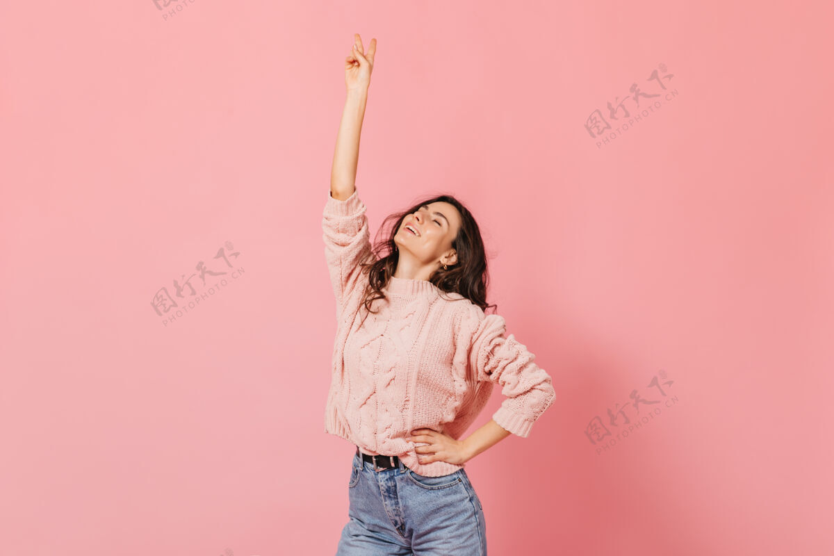 毛衣身穿粉色毛衣的苗条黑发女孩举手摆姿势 在孤立的背景上显示和平标志点粉色背景女孩