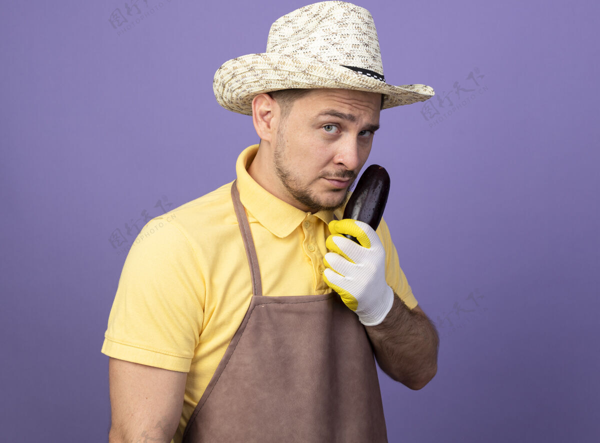 看年轻的园丁穿着连体衣 戴着帽子 戴着工作手套 手里拿着茄子 严肃的脸站在紫色的墙上看着前方花园工人连身衣