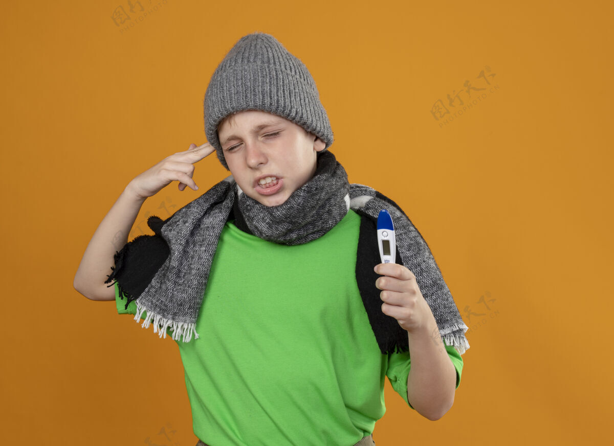 帽子生病的小男孩穿着绿色t恤 戴着暖和的围巾 戴着帽子 显示体温计 感觉不舒服 生病 不高兴 在站在橘色墙上的寺庙附近做着手枪手势手枪围巾体温计
