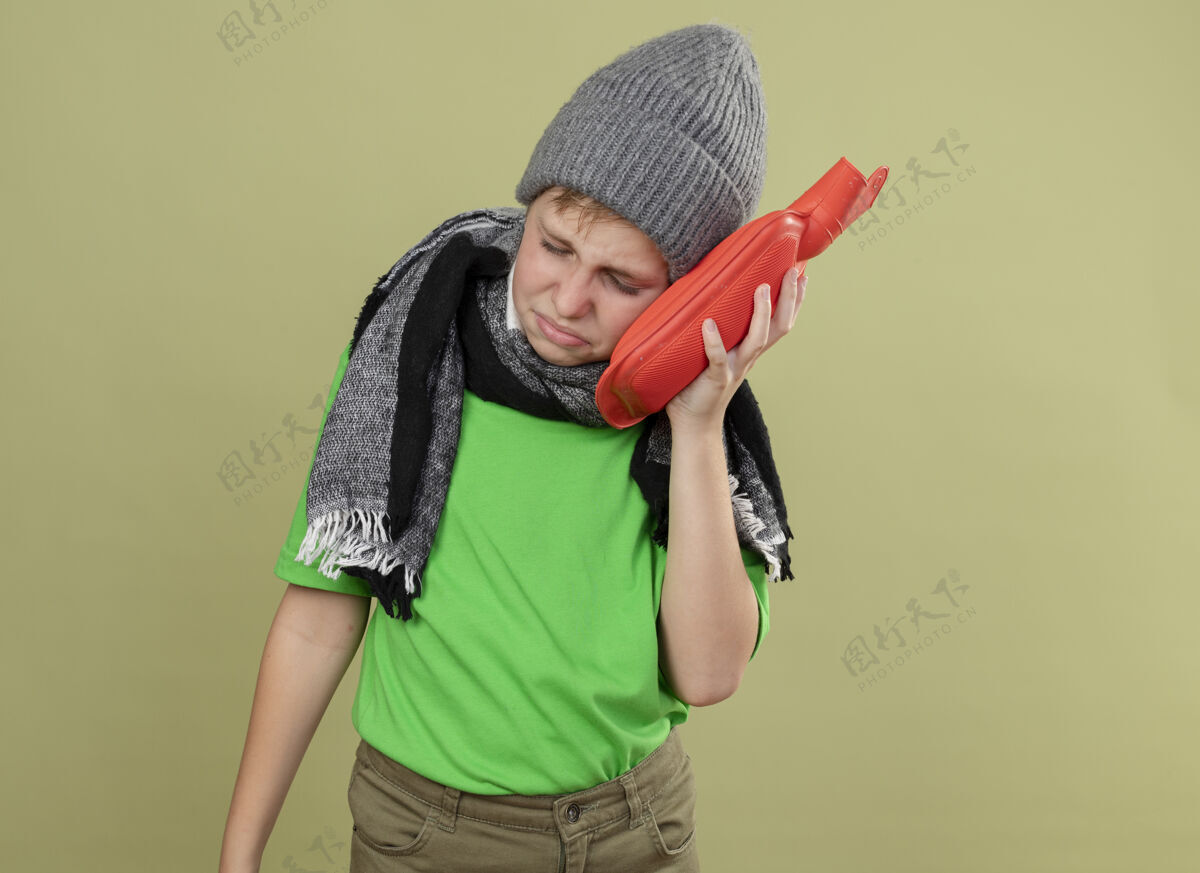 少生病的小男孩穿着绿色t恤 戴着暖和的围巾和帽子 感觉不舒服 拿着水瓶取暖 把头靠在上面 生病了 不高兴地站在轻质的墙上生病保持光