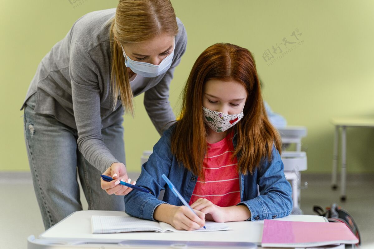 导师戴着医用口罩的女老师在课堂上帮助小女孩的正面图面罩流行病新常态