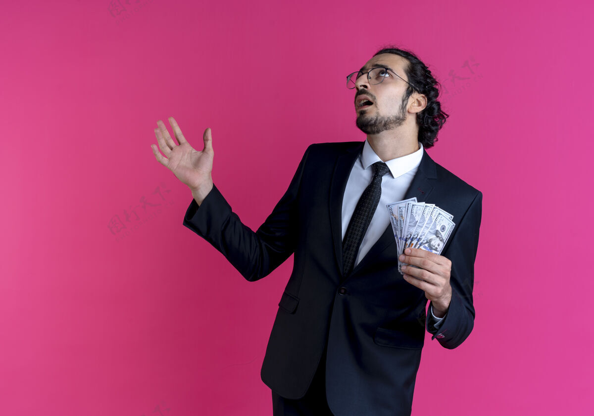 眼镜一个穿着黑色西装 戴着眼镜 拿着现金的商人 困惑地抬起头 伸出手臂站在粉红色的墙上男人西装姿势