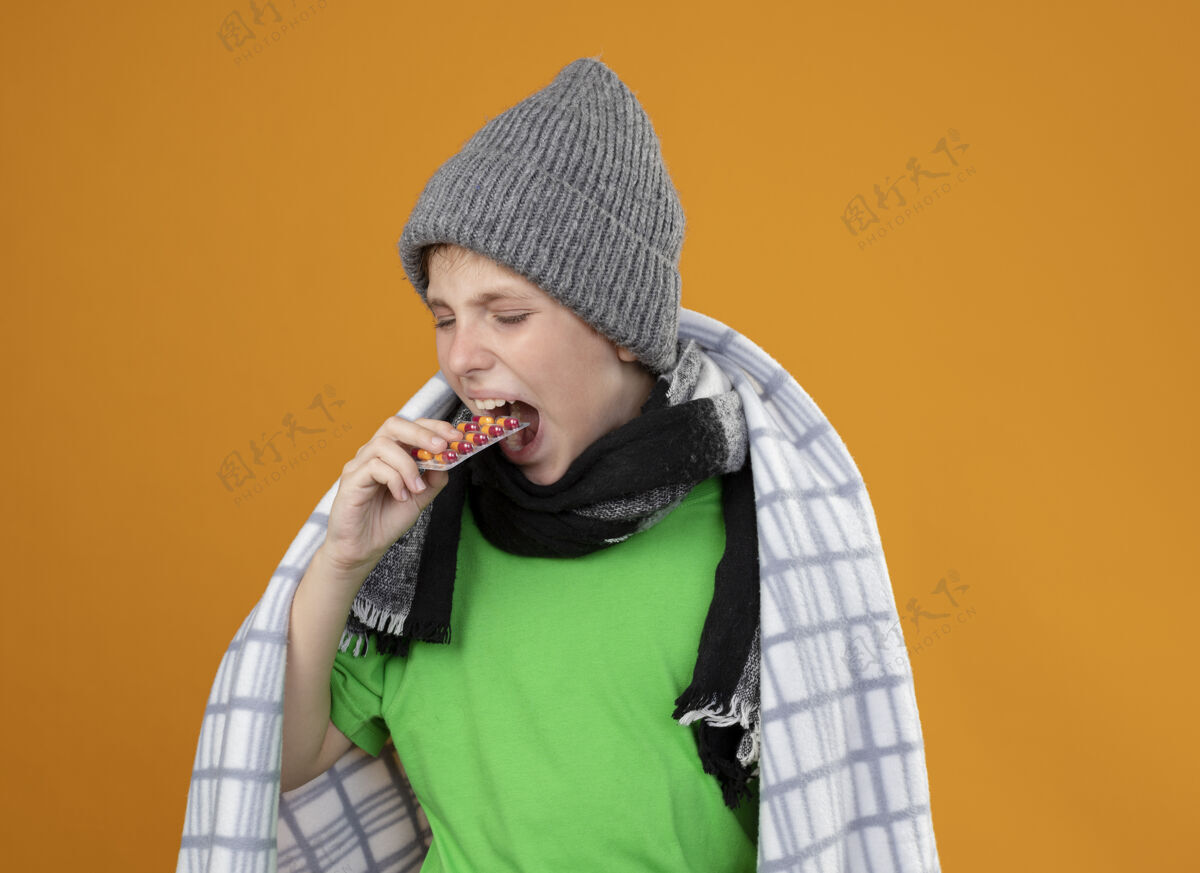 水泡生病的小男孩戴着暖和的帽子 围着围巾裹着毯子 把泡在嘴里的药丸放进橘子墙上站着温暖立场小