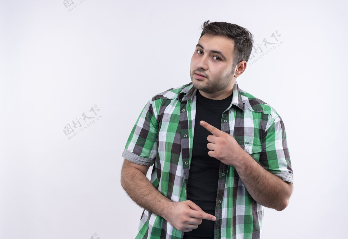 假日身穿格子衬衫的年轻旅行家站在白墙上 用手指着侧面 表情自信严肃表情市民自信