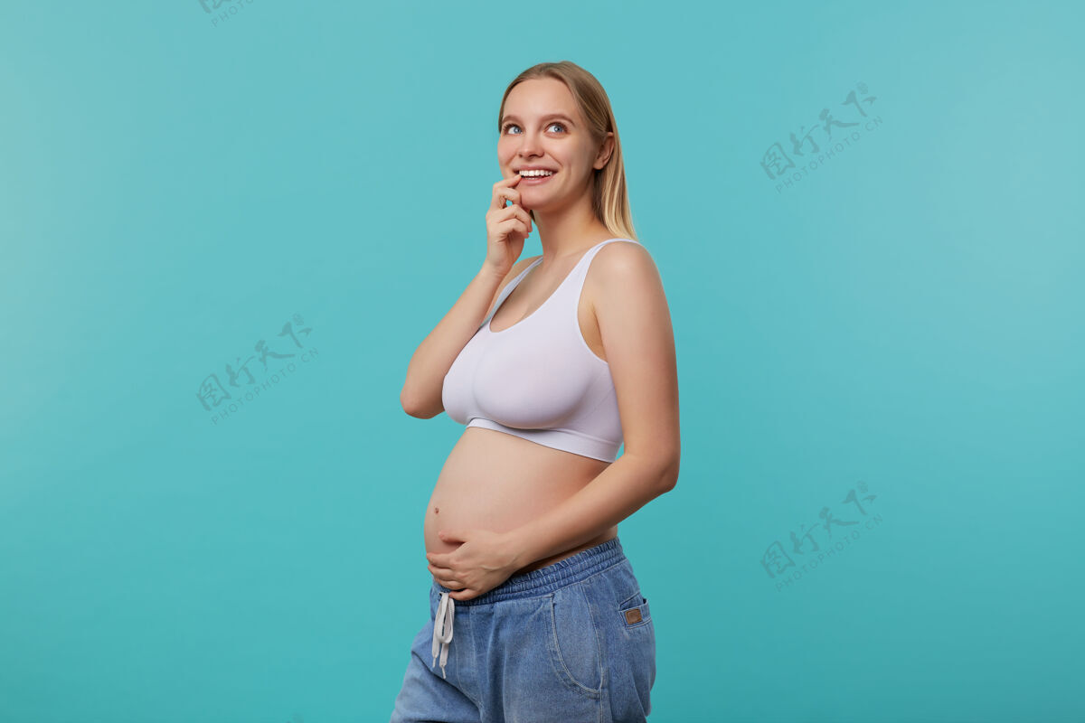 未来一张室内照片 年轻的怀孕的金发长发女人抱着她的肚子 面带愉快的微笑沉思地向上看 站在蓝色的背景上自然女性微笑