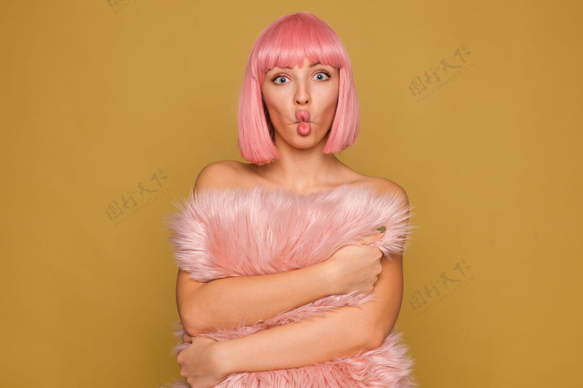 芥末有趣的镜头年轻迷人的粉红色头发的女性与短时髦的发型 使脸 而在芥末墙上摆姿势 可爱的毛茸茸的枕头在她的手中看女士邦