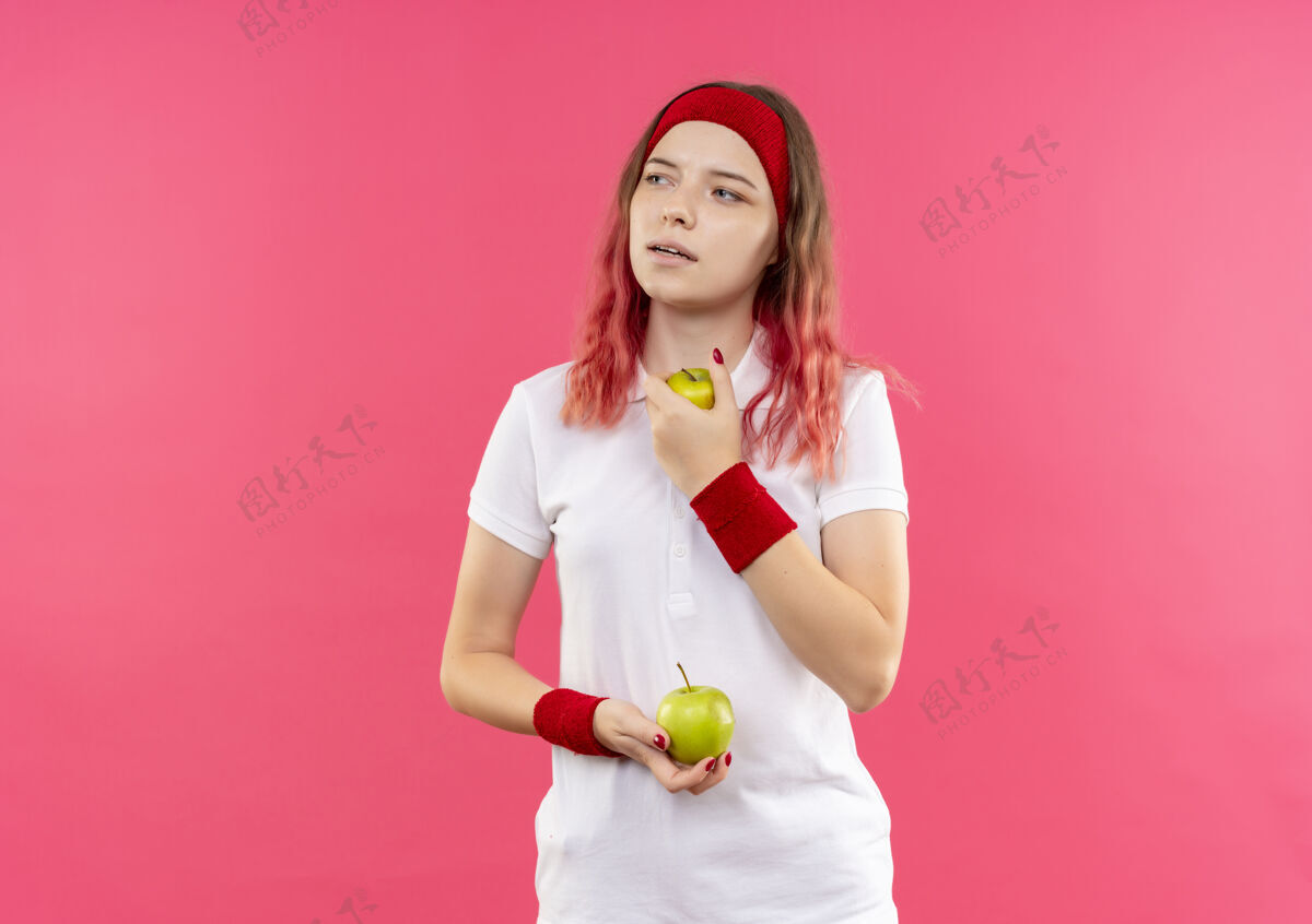 市民戴着头巾的年轻的运动型女人拿着两个苹果站在粉红色的墙上 带着沉思的表情望向一边一旁姿势抱着