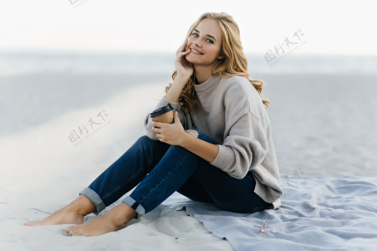 女性穿着牛仔裤的快乐女人坐在沙滩上喝着咖啡迷人的盲人女人在秋天的沙滩上摆姿势年轻女人周末