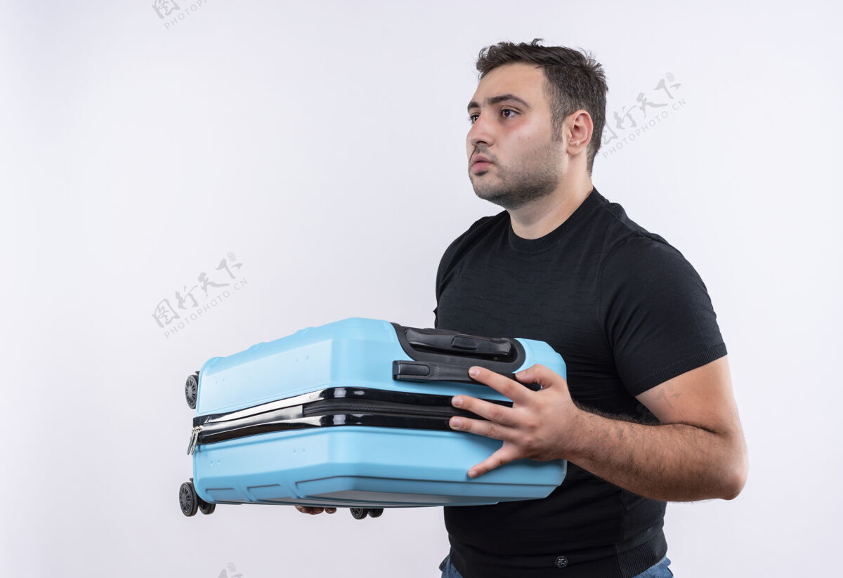 旅行身穿黑色t恤的年轻旅行家拿着手提箱 表情严肃地站在白色的墙上向一边看人旅行者年轻人