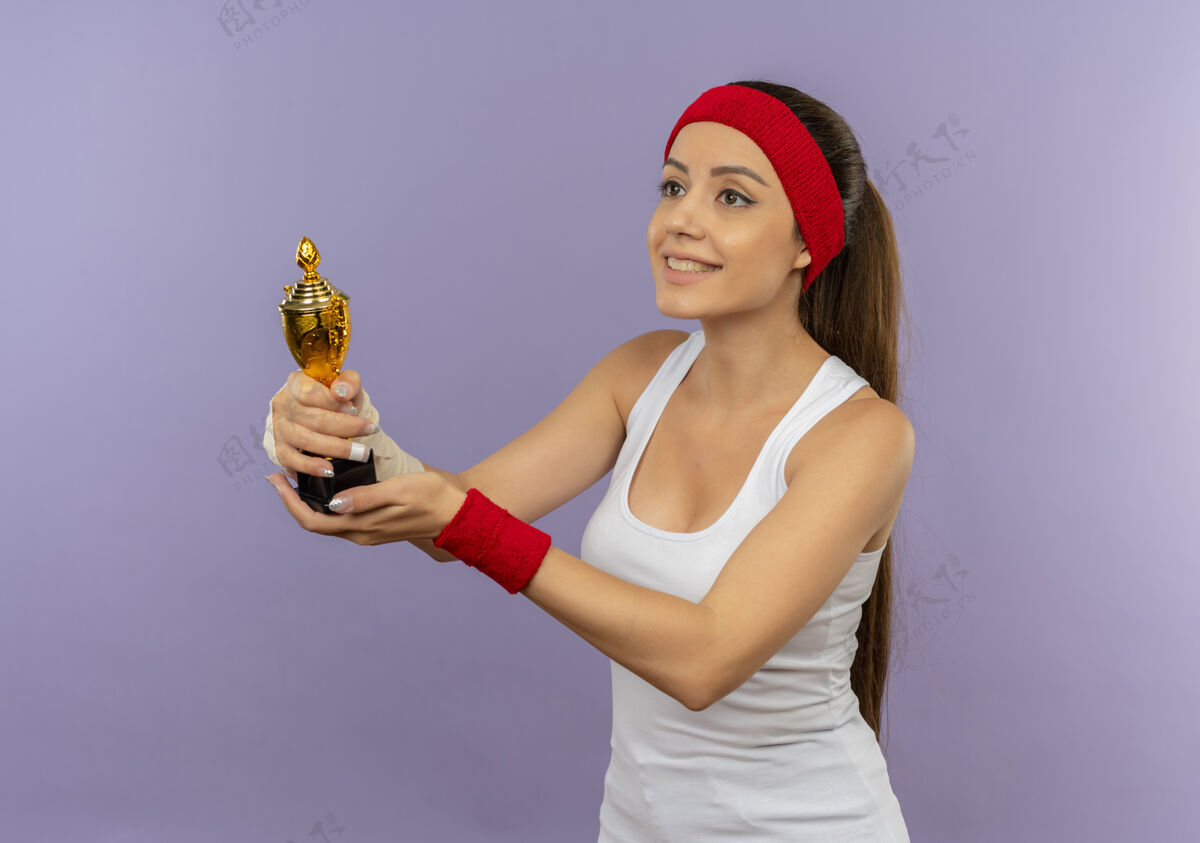 头带穿着运动服的年轻健身女 头上戴着头巾 手里拿着奖杯 一边看着灰色的墙上站着一张幸福的脸站着市民健身