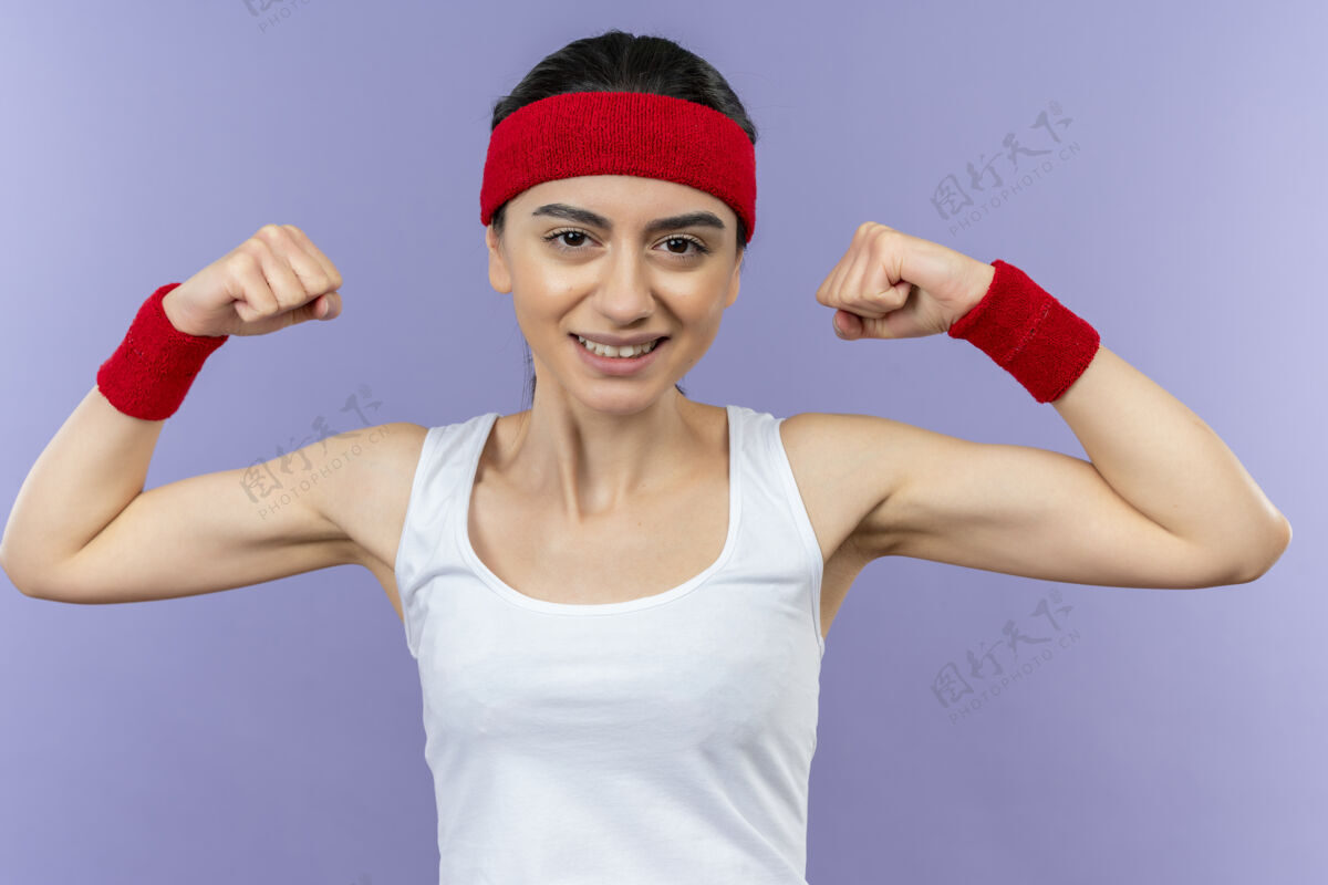 站立身穿运动服的年轻健身女士 头戴花环 微笑着 高举拳头 站在紫色的墙壁上 展现出快乐和积极的二头肌运动市民举起