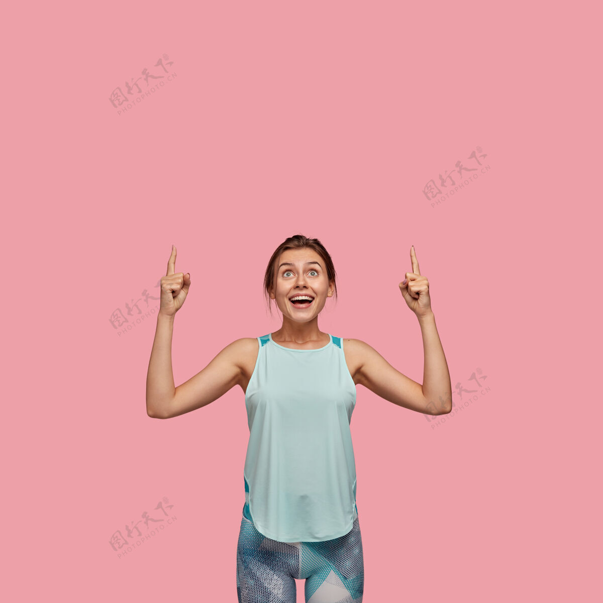 时尚积极的欧洲女人 表情愉快 食指朝上 穿着休闲背心和紧身裤 模特们站在粉红色的墙上广告概念看天花板大笑广告休闲