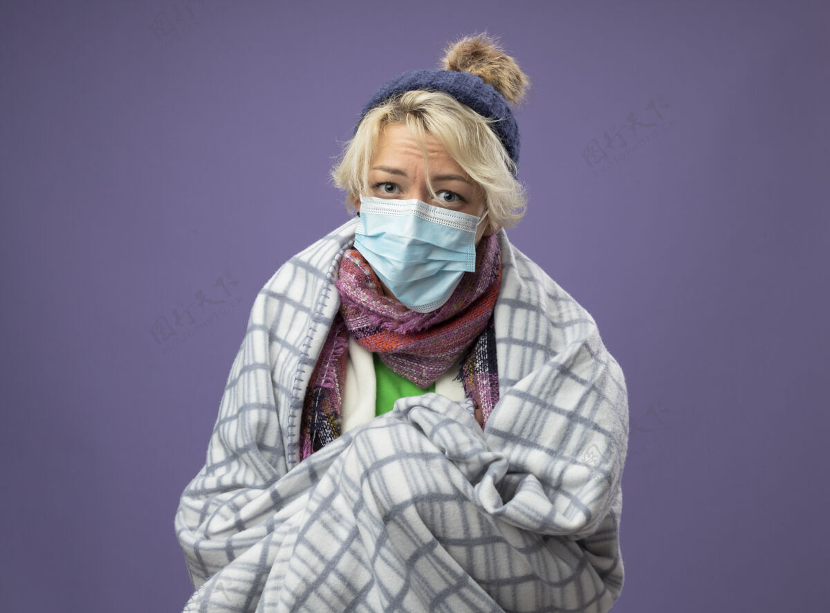 围巾生病的不健康的女人 短发 戴着暖和的帽子 戴着围巾 戴着防护面罩 裹着毛毯 感觉不舒服 看着相机 心情不好 背景是紫色的帽子温暖感觉